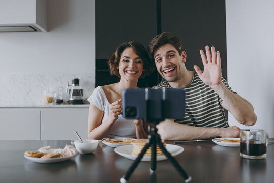 Young couple two woman man in casual clothes sit by table eat pancake talk video call mobile cell phone drink waving hand drink coffee cook food in light kitchen at home together Healthy diet concept