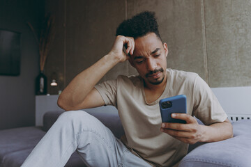 Pensive thoughtful sad young african american man wearing beige t-shirt sit on grey sofa indoors apartment using mobile cell phone prop up forehead think on weekends stay home. Tattoo translate fun.