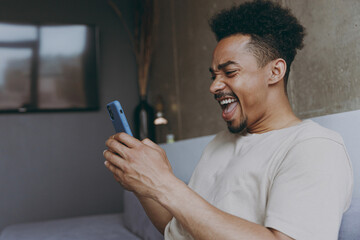 Side view laughing young african american man wear beige t-shirt sit on grey sofa indoor apartment hold mobile cell phone mocking friends joking watching memes internet rest on weekends stay at home - obrazy, fototapety, plakaty