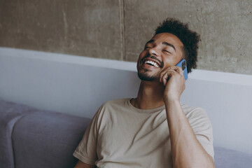 Close up joyful satisfied excited young african american man 20s wearing beige t-shirt sit on grey...