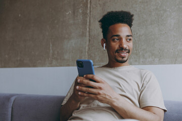 Young african american man in casual beige t-shirt sit on grey sofa indoors apartment use air pods mobile cel phone listen to music audiobook podcast look aside resting on weekends staying at home