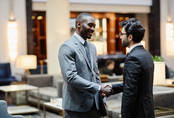 Side view portrait of two successful businessmen shaking hands while standing in hotel lobby, copy space