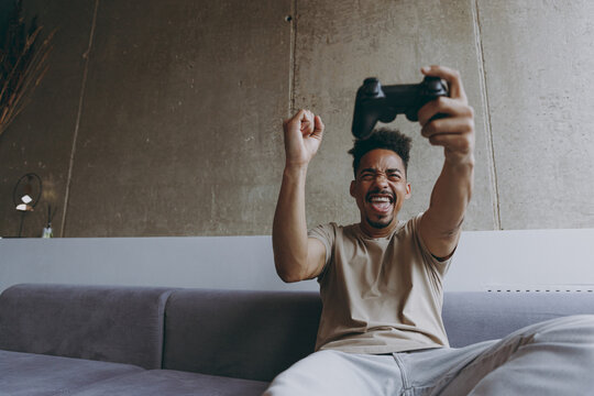 Bottom view epressive addicted young african american man in beige t-shirt sit on grey sofa indoors apartment play pc game by joystick console do winner gesture clench fist rest on weekends stay home