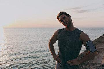 Fototapeta premium Young strong athletic toned fit sportsman man in sport clothes earphones mobile cell phone warm up train do head tilts at sunrise sun dawn over sea beach outdoor on pier seaside in summer day morning