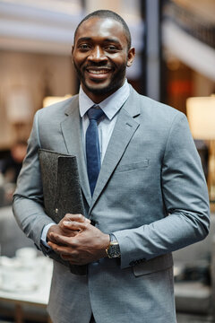 Vertical waist up portrait of successful African-American businessman smiling at camera while standing in hotel lobby