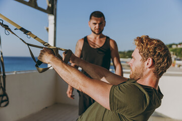 Young profile sporty strong athletic toned fit sportsman man in sport clothes warm up suspended training TRX work out with trainer at sunrise sun over sea beach seaside outdoor in summer day morning