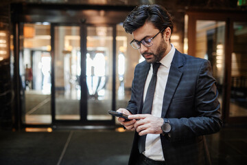 Waist up portrait of successful Middle-Eastern businessman using smartphone while standing in hotel lobby, copy space