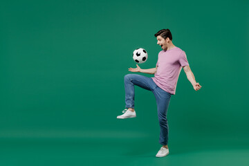 Fototapeta na wymiar Full size body length young fun man fan wears basic pink t-shirt cheer up support football sport team juggling soccer ball on knee watch tv live stream isolated on dark green color background studio.