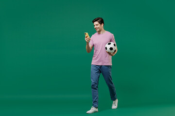 Naklejka na ściany i meble Full size body length fun young man fan in pink t-shirt cheer up football sport team hold hand soccer ball mobile cell phone watch tv live stream scream isolated on dark green color background studio.