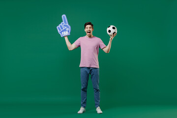 Fototapeta na wymiar Full size body length young man fan wear basic pink t-shirt cheer up support football sport team hold in hand soccer ball watch tv live stream scream isolated on dark green color background studio
