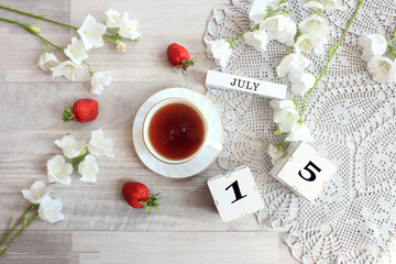 Calendar for July 15 : cubes with the number 15, the name of the month of July in English, a cup of tea on a gray openwork napkin, scattered flowers of bluebells and strawberries, top view