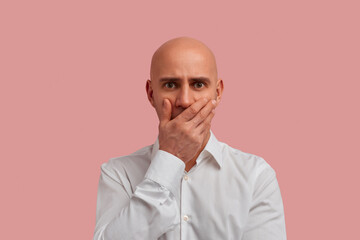 Ouch. I really said it. Close up portrait of bald man with bristle in white shirt cover open mouth by hand palm, look at camera with disappointments eyes, isolated over pink background.