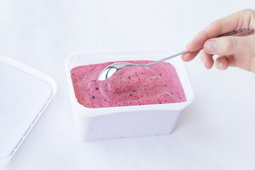 Berry black currant ice cream pink color cream texture in a rectangular white container with an open lid and metal silver spoon in hand on a white background. Top view. Eating concept