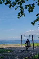 An sad adult guy, a man in a cap and with a backpack sits on a swing and sways among the green trees against the background of the blue sea and sky. Back view