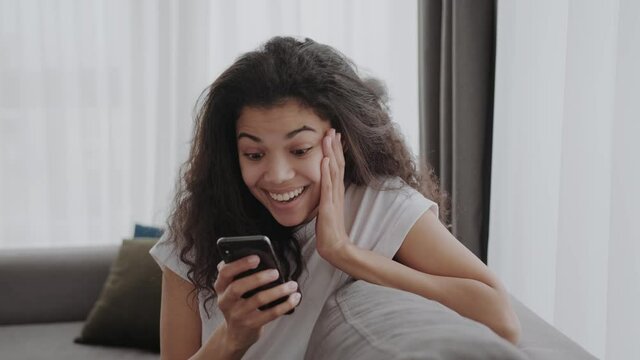 black young woman looks into a smartphone and is pleasantly surprised and happy