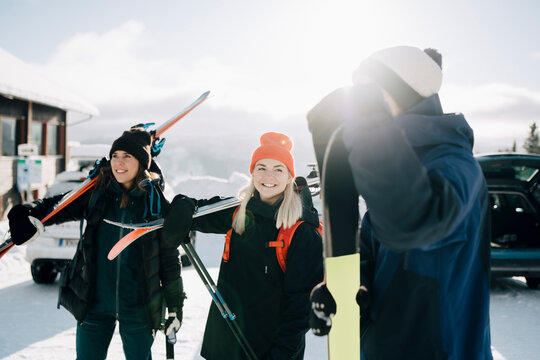 Leende v&auml;nner st&aring;r med skidor p&aring; parkeringsplats under solig dag