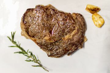 Grilled ribeye beef steak served on marble board with rosemary and grilled garlic; top view.