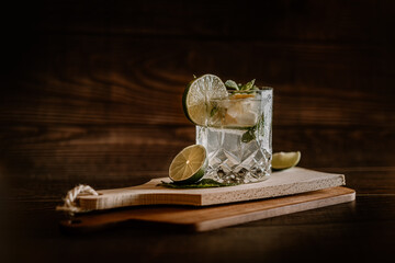 Moody dark photography of a cold drink, a glass of mojito with mint leaf, ice, lemon and sugar on a wooden table with a vintage style.
