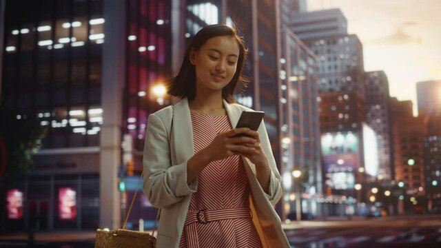 Portrait of an Attractive Japanese Female Wearing Smart Casual Clothes and Using Smartphone in a City at Sunset. Stylish Woman Connecting with People Online, Messaging and Browsing Internet.
