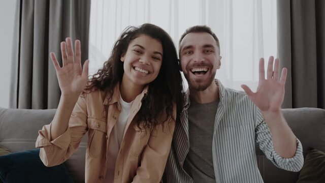 Portrait of happy multicultural couple sitting on couch at home, waving hands at camera, friendly interracial spouses greeting somebody, gesturing hello while relaxing on sofa.