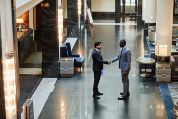 Wide angle view at two business partners shaking hands while standing in luxurious hotel lobby,...