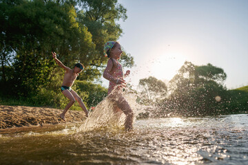 Happy children run from the shore into the water. Summer children's vacation on shore of a lake or river. boy and girl jump into water, swim and splash around at sunset. Active holidays. Dynamic image