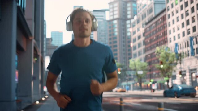 Handsome Young Man is Jogging on a Street of a Big City Center. Male is Running in Blue T-Shirt, Wearing Wireless Headphones with His Favourite Music Playing. Urban Training Workout in the Morning.