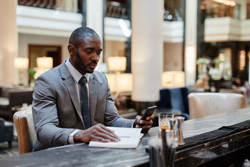 Portrait of successful African-American businessman using smartphone while relaxing at bar in hotel...