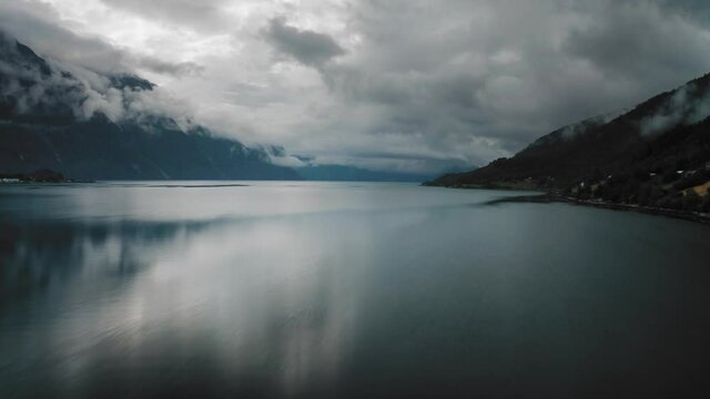 Gloomy heavy clouds are leisurely spreading above Romsdalenfjord, Norway, while capturing a deep blue fjord surface from the air. A thick dark forest covers the hills, where a few houses popped up.