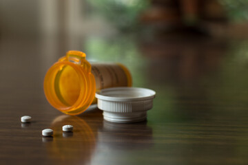 Bottle of prescription pills in an orange plastic bottle with white lid; three small white pills lay on table with out of focus dinging room in background - obrazy, fototapety, plakaty