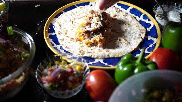 slow motion shot of cheese being dropped on top of a flat bread for a taco tortilla fajita with the sides of fresh vegetables like tomatos bell peppers chillies and rice in dramatic light