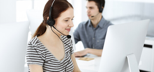 Casual dressed young woman using headset and computer while talking with customers online. Group of operators at work. Call center, business concept