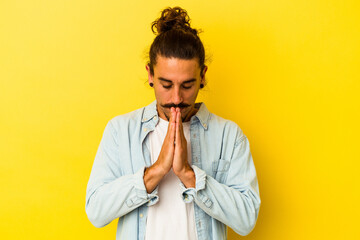 Young caucasian man with long hair isolated on yellow background praying, showing devotion, religious person looking for divine inspiration.