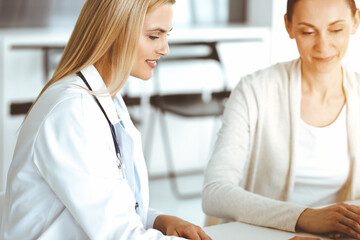 Fototapeta premium Woman-doctor at work in sunny hospital is happy to consult female patient. Blonde physician checks medical history record and exam results while using clipboard