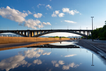 The sky and the Communal Bridge are reflected in a rain puddle. Urban landscape of the city of Krasnoyarsk on a summer day. City embankment of the Yenisei River