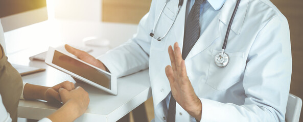 Doctor and patient discussing medical exam results while sitting at the desk in sunny clinic, close-up. Male physician using tablet computer for filling up medication history record.