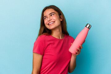 Young caucasian woman holding a water bottle isolated on blue background looks aside smiling, cheerful and pleasant. - Powered by Adobe