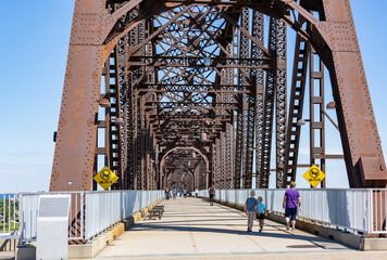 Entrance to a pedestrian bridge over the Ohio River in Louisville, Kentucky on a sunny day with a few people on the bridge.