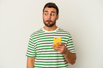 Young caucasian man holding a mobile phone isolated on white background confused, feels doubtful and unsure.