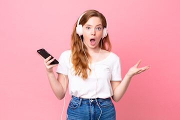 pretty red head woman feeling extremely shocked and surprised with headphones and smartphone