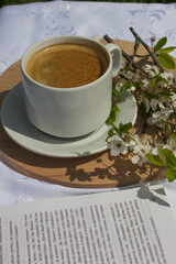 A cup of coffee, a book and blooming cherry branches on a white tablecloth. White sakura. Atmosphere, comfort, rest