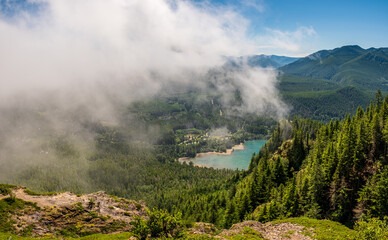 A fog cover hides a forested valley and blue lake below