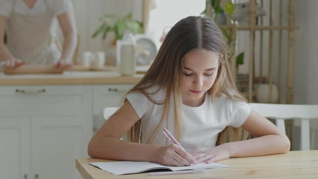Teenager girl child schoolgirl pupil sitting at home at table in kitchen doing homework writing exercise math study learning indoor homeschooling, adult mother cooks kneads dough using rolling pin