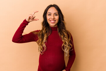 Young mexican pregnant woman isolated on beige background holding something little with forefingers, smiling and confident.