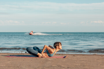 a young attractive woman, of Asian appearance, practicing yoga, performs a stretching exercise, on the beach. bhekasana, mandukasana, frog pose