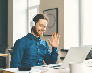 Young happy man office worker in headphones waving at webcam on laptop and smiling during video...