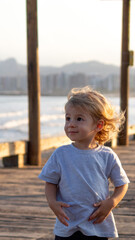 CRIANÇA LOIRA SOZINHA EM DECK, COM PRAIA NO FUNDO. 