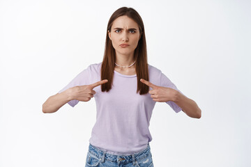 Grumpy young woman looks disappointed and points at herself, disagree, complain about unfait decision, standing displeased and frustrated against white background, what about me