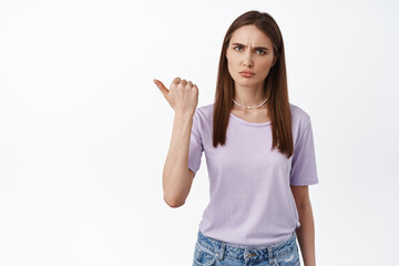 Displeased young woman pointing left and look skeptical, disbelief about product quality, frowning and grimacing judgemental, white background