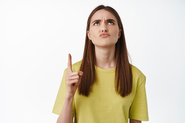 Portrait of disappointed girl, frowning and sulking, pointing finger up at smth strange,...